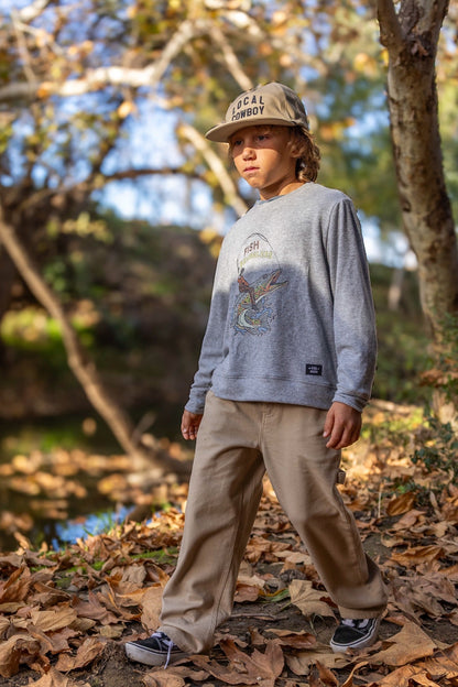 A child standing outdoors in a wooded area with fallen leaves, wearing a heather gray graphic fish wrangler sweatshirt, beige pants, a beige cap, and dark sneakers.