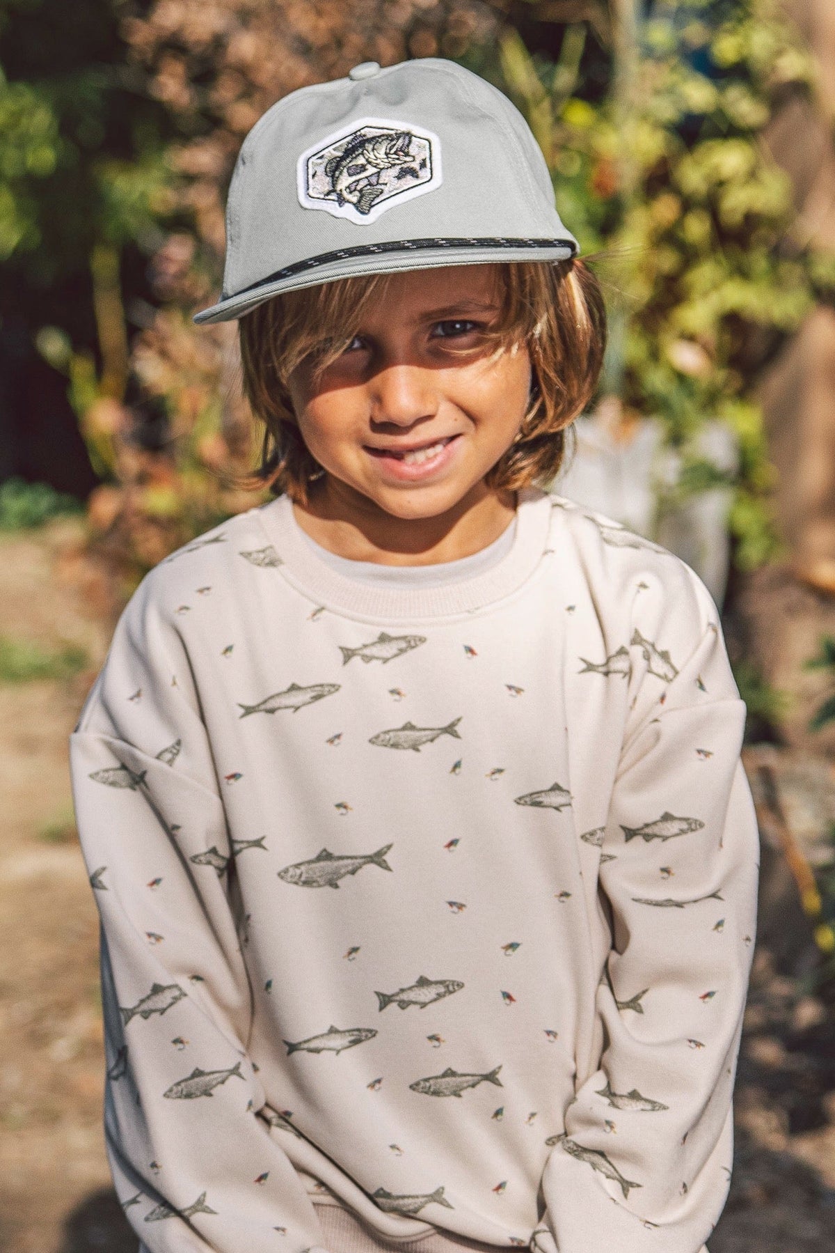 A young child wearing a light gray gone fishing snapback cap with a fish patch and a beige sweatshirt covered in small shark prints, standing outdoors in front of a blurred natural background.