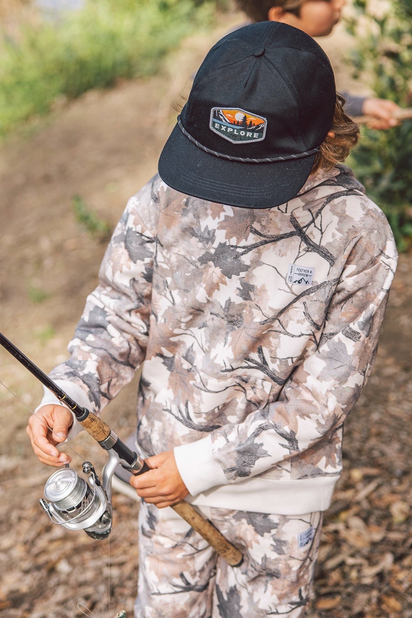 A child wearing a camouflage hunting outfit and a black Explore cap, holding a fishing rod outdoors on a wooded path.