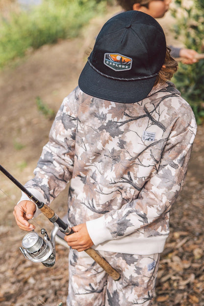 A child wearing a camouflage hunting outfit and a black Explore cap, holding a fishing rod outdoors on a wooded path.