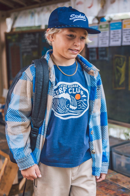 A child wearing a navy blue baseball cap with 'Stoked' embroidered on it, a blue graphic t-shirt, a blue and beige plaid flannel shirt, and khaki pants, standing in a rustic shop environment.