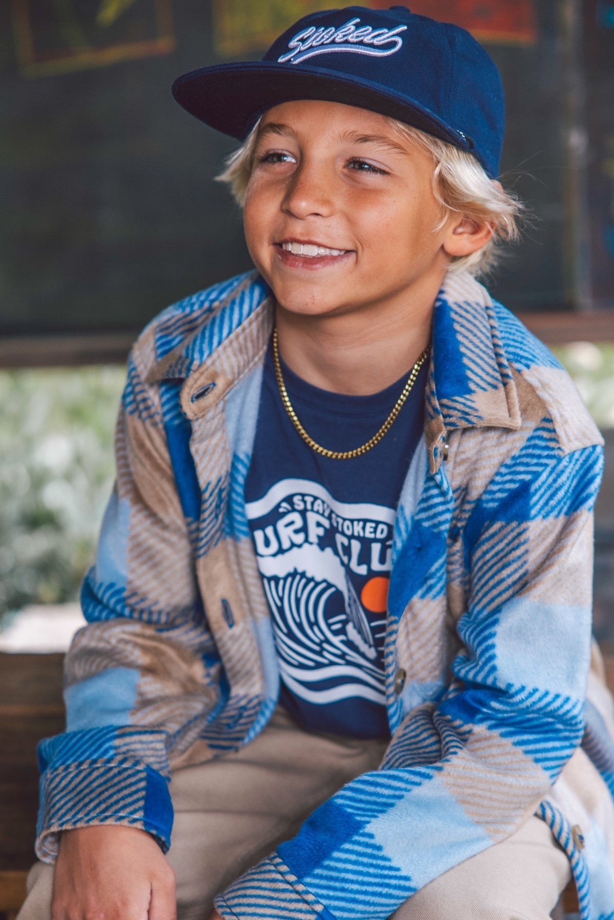 A smiling young boy wearing a blue and beige plaid flannel shirt over a navy t-shirt with a wave graphic, paired with a navy baseball cap with Stoked embroidered on it, sitting in a rustic outdoor setting.