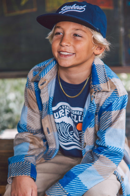A smiling young boy wearing a blue and beige plaid flannel shirt over a navy t-shirt with a wave graphic, paired with a navy baseball cap with Stoked embroidered on it, sitting in a rustic outdoor setting.