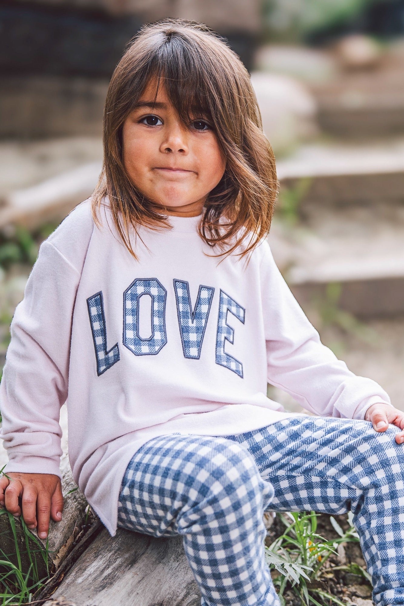 A young girl with brown hair sits on a log outdoors, wearing a light pink sweatshirt with 'LOVE' printed in blue-and-white gingham letters, paired with matching crown blue-and-white gingham pants.