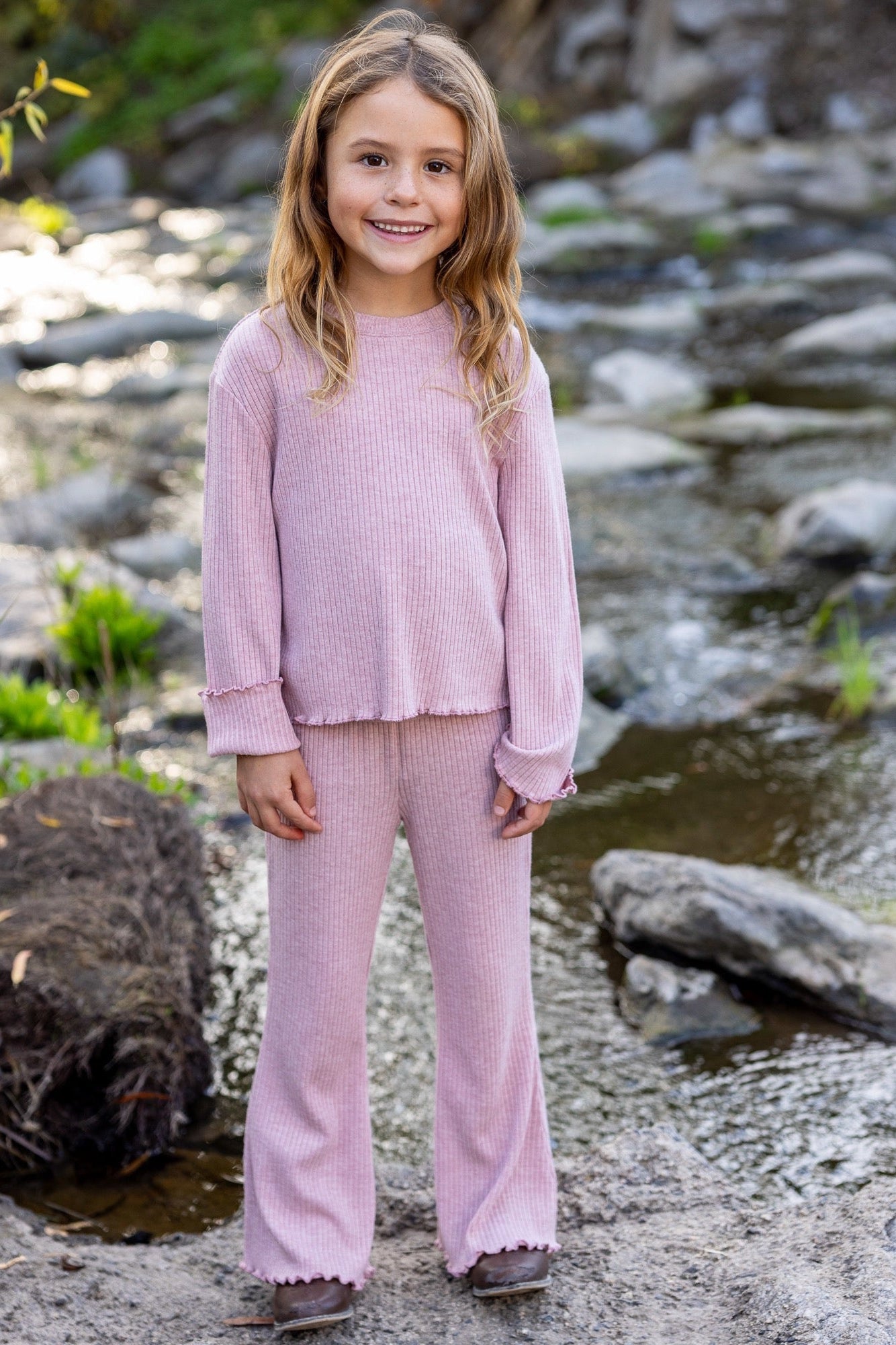 A young girl stands outdoors in front of a rocky stream wearing a matching light rose pink ribbed knit lounge set consisting of a long-sleeve top and meadow bell bottom wide-leg pants.