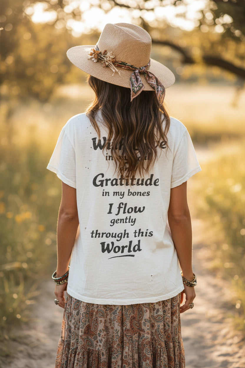 A woman viewed from behind wearing a cream worn weathered art graphic t-shirt with black text, a beige straw hat with a patterned ribbon, and a brown patterned skirt, standing on a sunlit path outdoors.