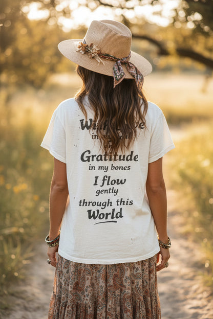 A woman viewed from behind wearing a cream worn weathered art graphic t-shirt with black text, a beige straw hat with a patterned ribbon, and a brown patterned skirt, standing on a sunlit path outdoors.