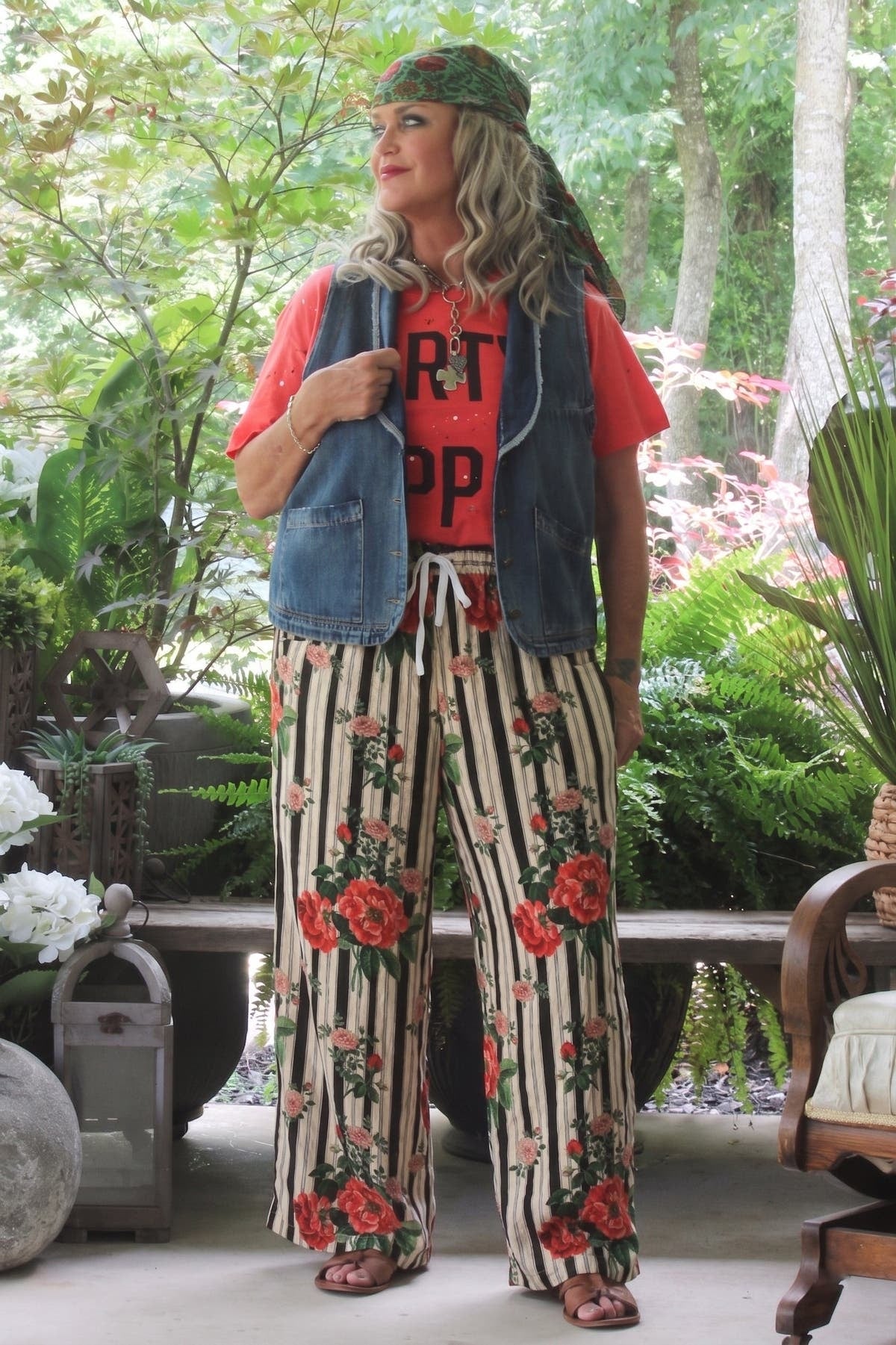 A woman stands on a patio wearing a red graphic t-shirt, a blue denim vest, wide-leg striped pants with a red rose floral print, brown sandals, and a green patterned cap. The background features lush green garden plants and trees.