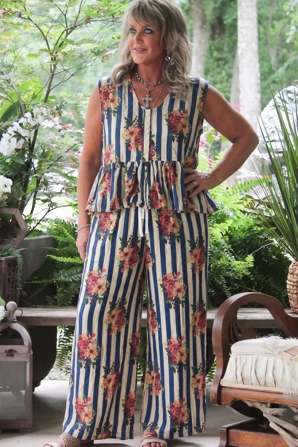 A woman modeling a sleeveless peplum starlight drift tank top with vertical blue and cream stripes and a floral print, standing on a patio with plants and furniture in the background.