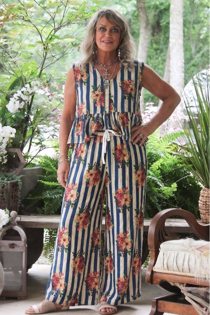 A woman modeling a sleeveless starlight drift matching set with vertical royal navy and cream stripes and a floral print, standing on a patio with plants and furniture in the background.