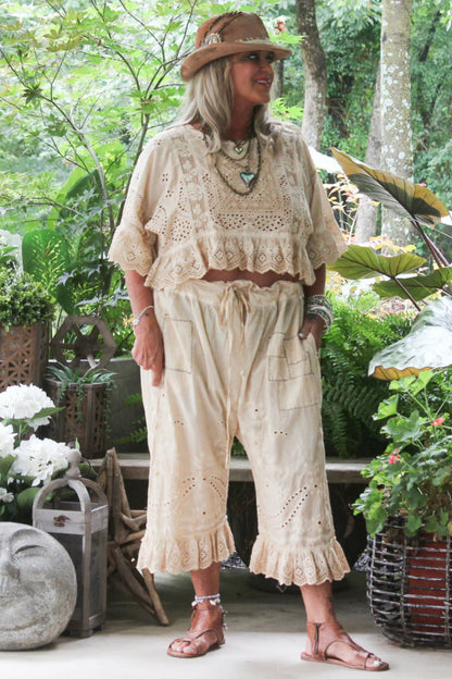 A woman wearing a bohemian-style oatmeal colored crochet two-piece set with a softly worn eyelet cropped top and drawstring pants, styled with a brown hat and sandals in a lush garden setting.