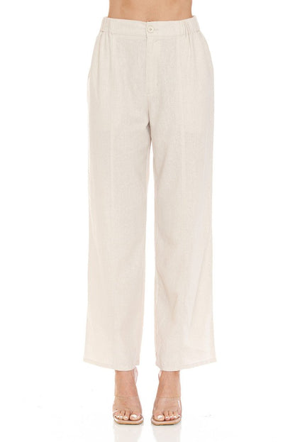 A model is wearing a pair of high-waisted, wide-leg cropped pants in a light cream or khaki linen-like fabric, paired with clear high-heeled sandals against a solid white background.