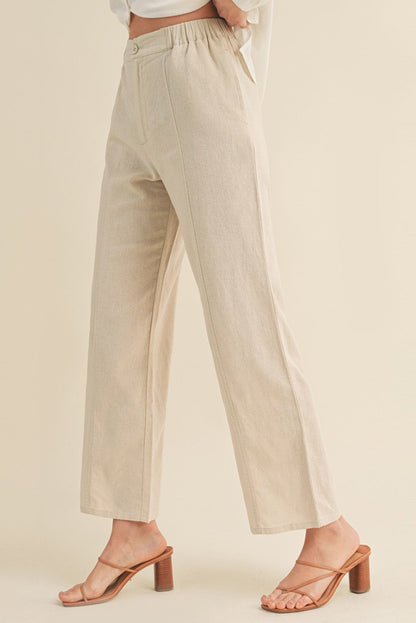 A model wearing high-waisted, wide-leg linen trousers in a light beige khaki color, paired with brown block-heel slide sandals, against a solid cream background.
