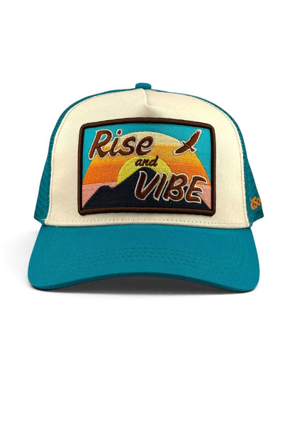 A teal and cream trucker hat with a rectangular embroidered patch on the front that reads 'Rise and Vibe' over a sunset and mountain design.