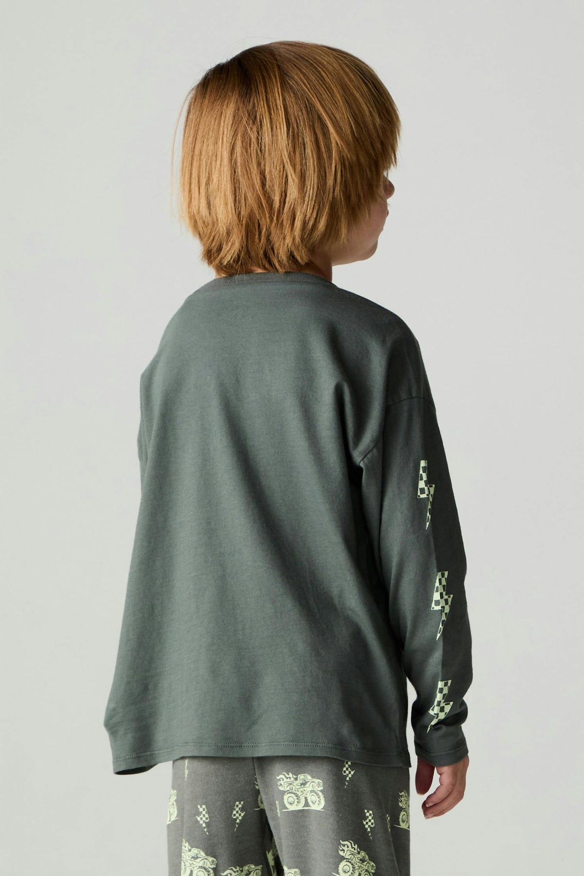 A child shown from the back wearing a dark safari green long-sleeve top with light green graphic patterns on the sleeves and gray pants with a matching green print, against a plain light gray studio background.