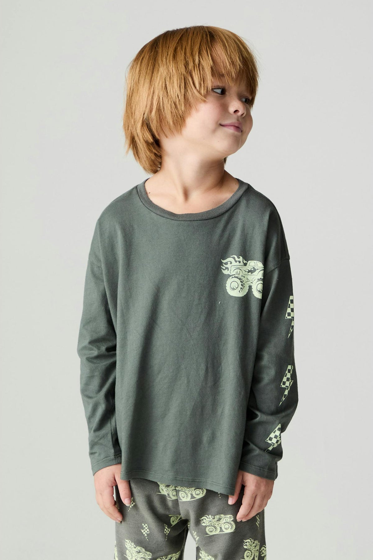 Boy wearing a safari green long sleeve tee with a monster truck graphic print on the front and down one arm paired with matching monster truck pants