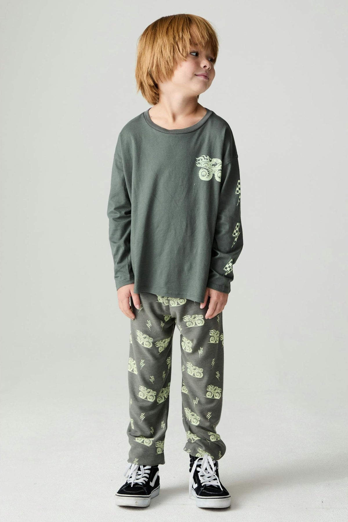 Boy wearing a safari green long sleeve tee with a monster truck graphic print on the front and down one arm paired with matching safari green monster truck pants