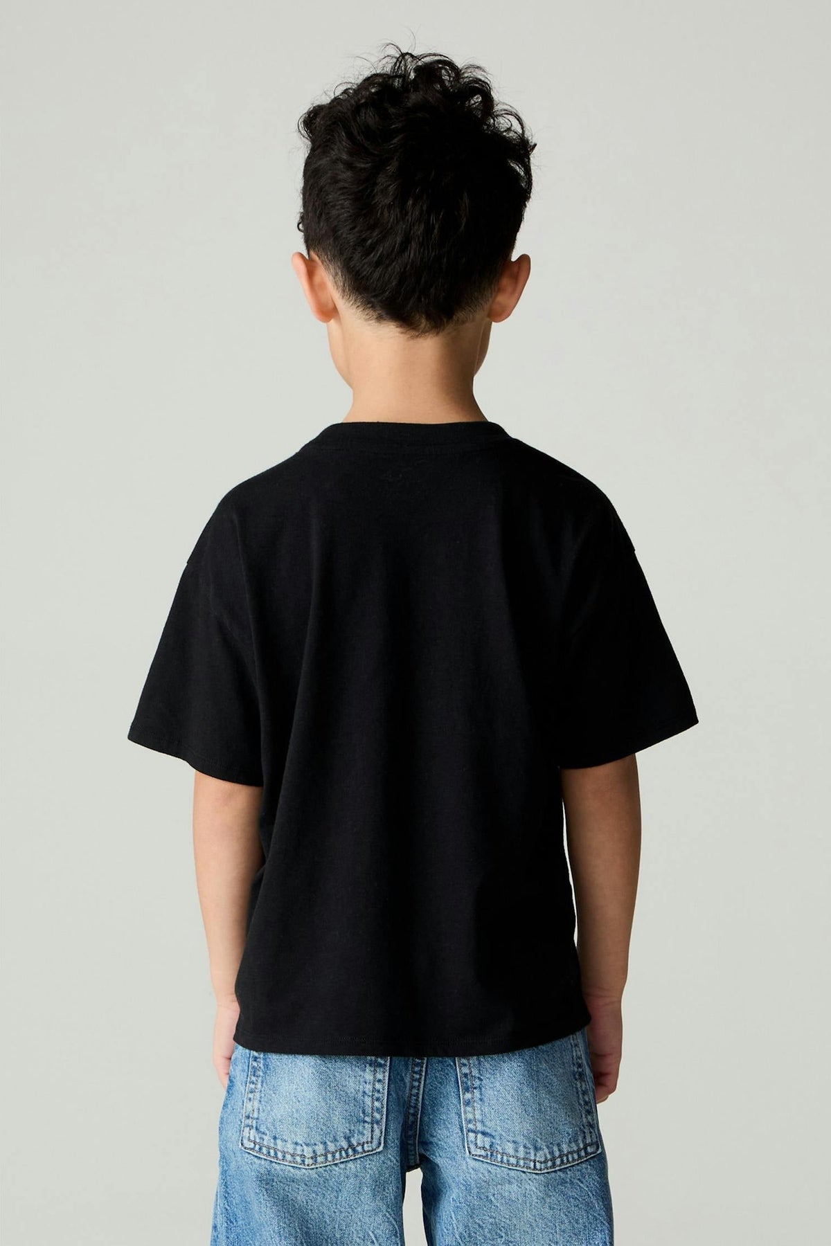 A child viewed from behind wearing a plain black short-sleeve t-shirt and blue jeans, standing against a light gray studio background.