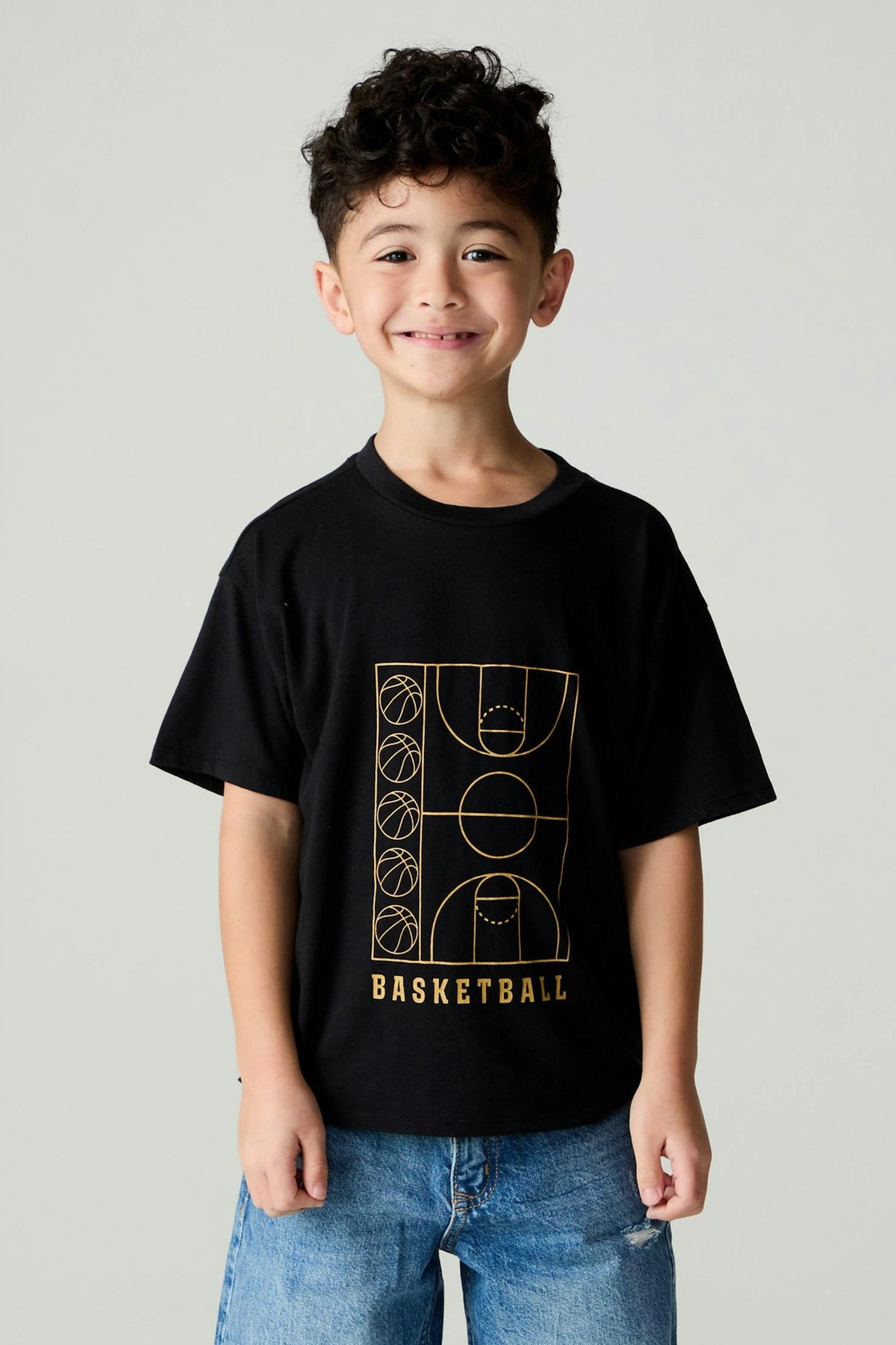 A young boy with curly dark hair smiling, wearing a black short-sleeve t-shirt with a gold basketball court graphic and the word 'BASKETBALL' on the front, paired with blue jeans, against a light gray studio background.