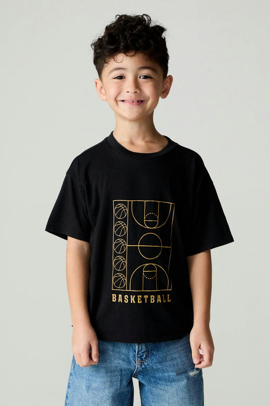 A young boy with curly dark hair smiling, wearing a black short-sleeve t-shirt with a gold basketball court graphic and the word 'BASKETBALL' on the front, paired with blue jeans, against a light gray studio background.