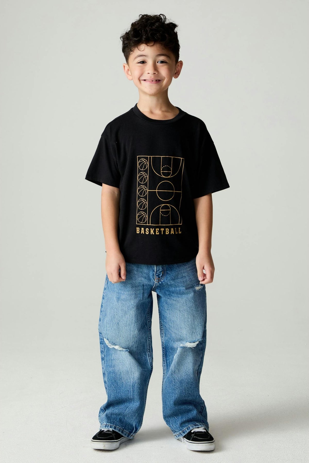 A young boy stands against a light gray studio background wearing a black short-sleeve t-shirt with a gold basketball court graphic and the word 'BASKETBALL' on the front, paired with wide-leg distressed blue jeans and black sneakers.