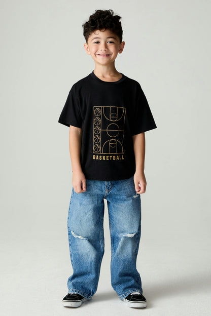 A young boy stands against a light gray studio background wearing a black short-sleeve t-shirt with a gold basketball court graphic and the word 'BASKETBALL' on the front, paired with wide-leg distressed blue jeans and black sneakers.