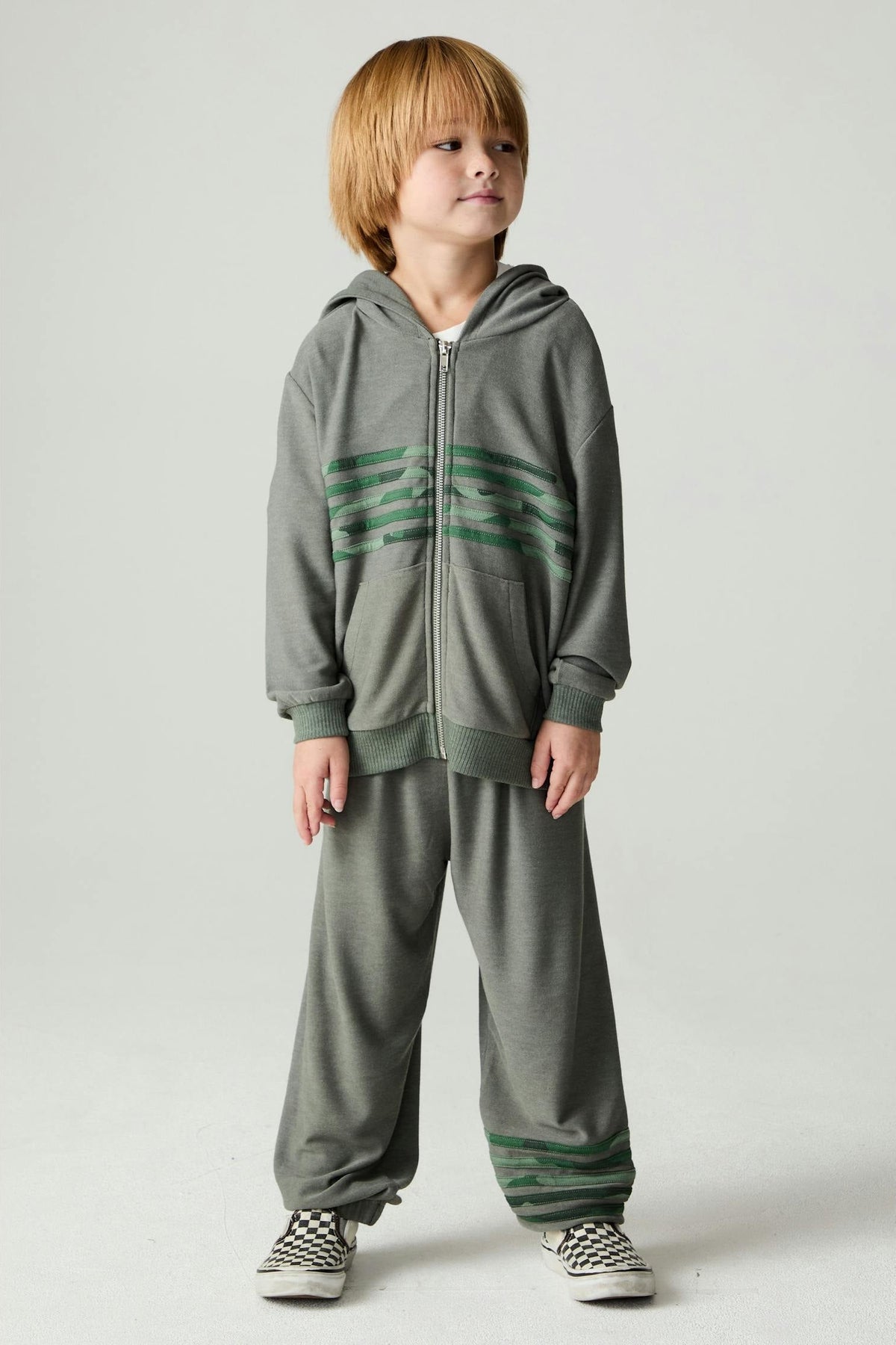 A young child wearing a matching safari green zip-up hoodie and sweatpants set with green horizontal camo stripes, standing against a light gray studio background.