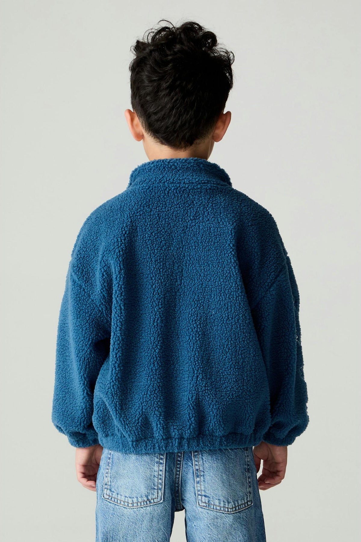 A child viewed from behind wearing a textured blue fleece sherpa pullover and light wash denim jeans, standing against a plain light gray studio background.