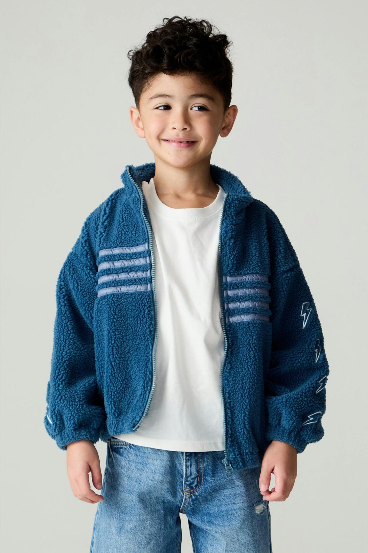 A young boy wearing a blue sherpa fleece zip-up jacket with white stripe details on the chest & lightning bolts on the arms styled over a white t-shirt and blue jeans, set against a plain light gray background.