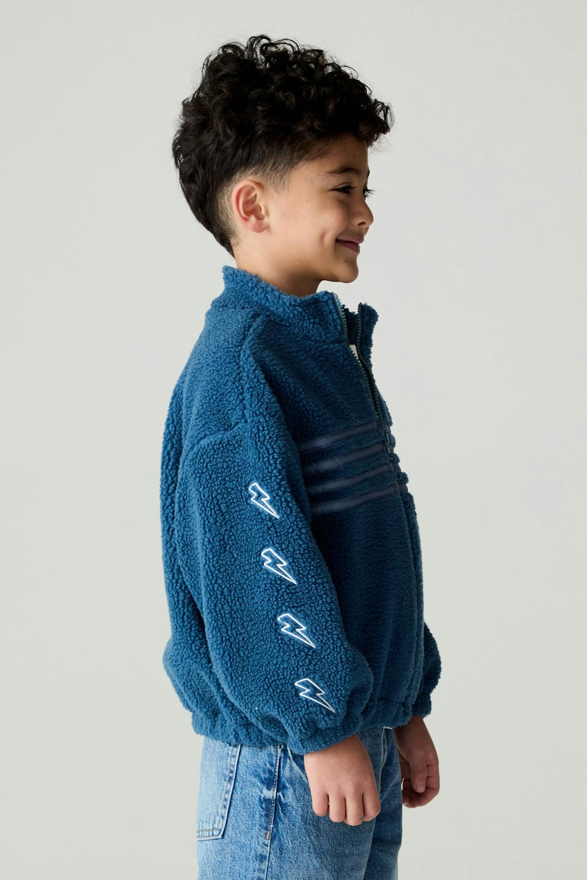 A young boy with curly dark hair is shown in profile wearing a blue fleece quarter-zip pullover sweater with white embroidered lightning bolt details on the sleeve, paired with blue jeans, against a plain light gray background.