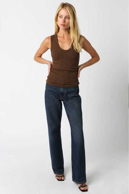 Woman wearing a brown thick strap tank top and dark blue jeans with brown sandals against a plain background 