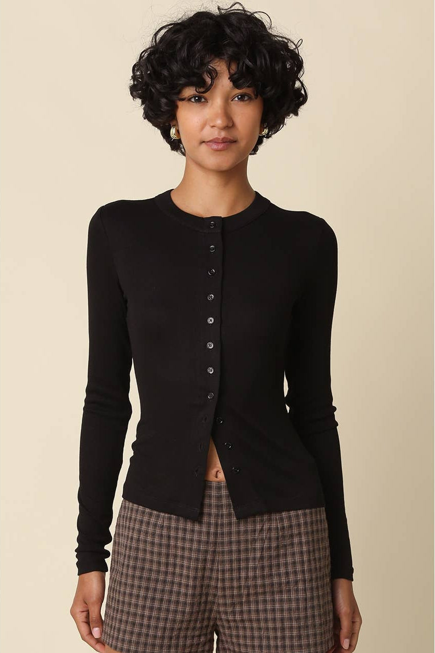 A model with short curly black hair wearing a black long-sleeve button-front cardigan and brown plaid pants, standing against a solid beige background.