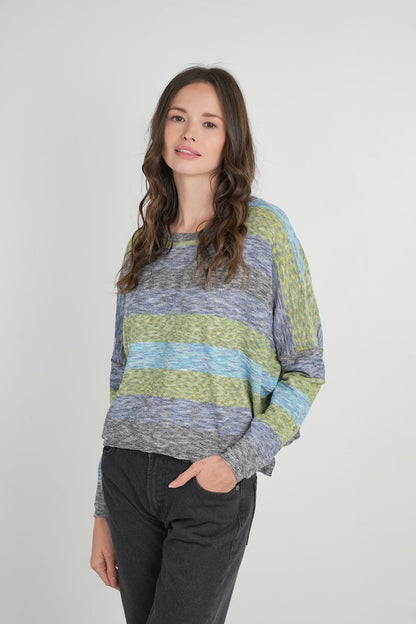 A woman modeling a long-sleeve knit sweater with horizontal stripes in heathered blue-gray, navy blue, sage green, and olive green, paired with dark gray jeans against a light gray studio background.