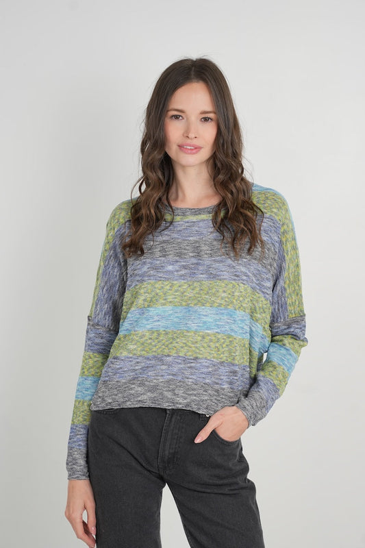 A woman modeling a long-sleeve knit sweater with horizontal stripes in gray, navy blue, and olive green, paired with dark gray jeans, against a plain light gray studio background.