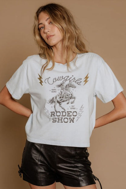 A woman modeling a white graphic t-shirt with a cowgirl rodeo theme, paired with black faux leather shorts, against a solid beige background.
