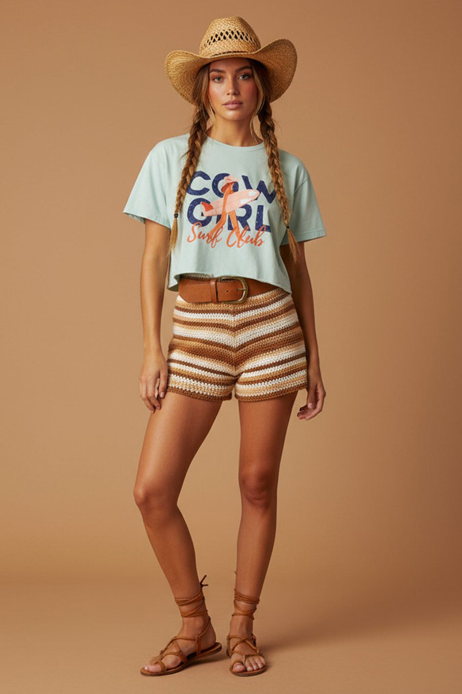 A woman modeling a light blue graphic t-shirt that reads 'COWGIRL Surf Club' in navy and orange, paired with brown and beige striped knit shorts, a straw cowboy hat, and brown lace-up sandals, set against a solid tan studio background.