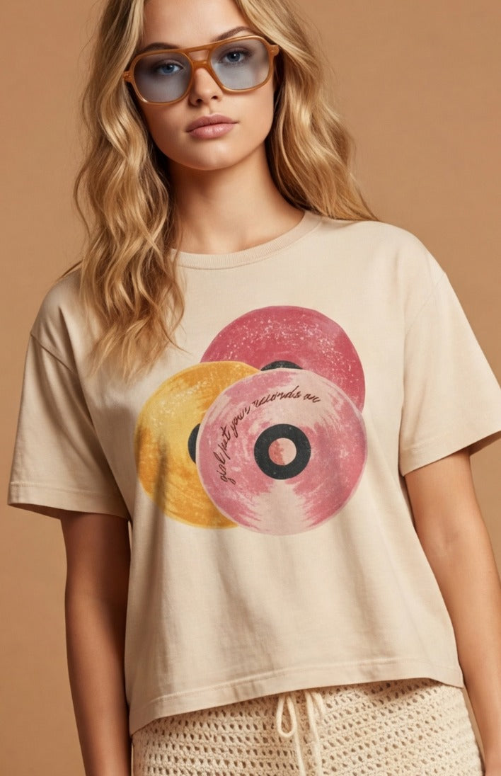 A woman wearing a beige cropped t-shirt with a graphic of overlapping pink and yellow vinyl records, set against a solid warm beige background.