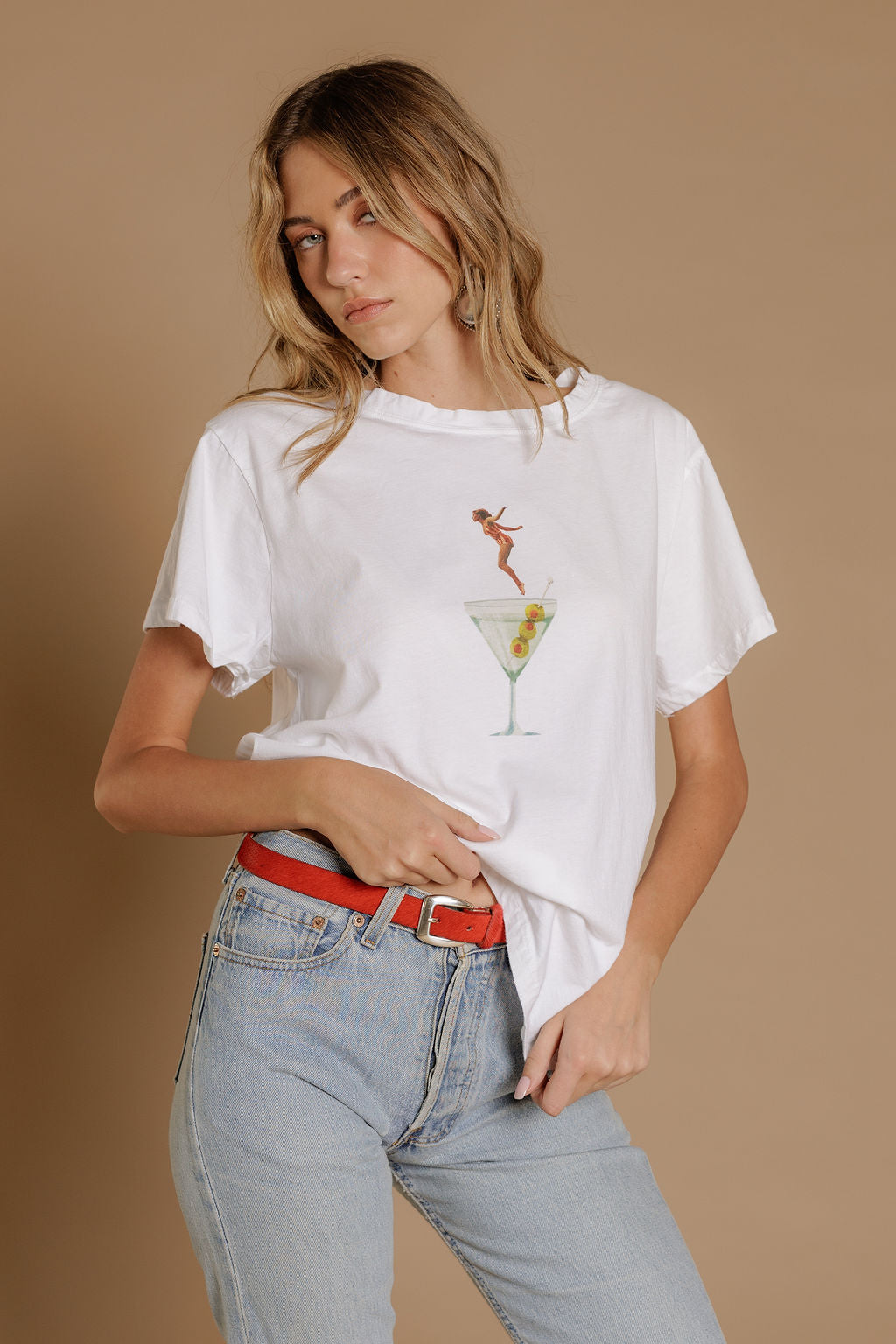 Woman wearing a white t-shirt with a graphic design of a woman jumping into a martini glass and light blue jeans on a beige background