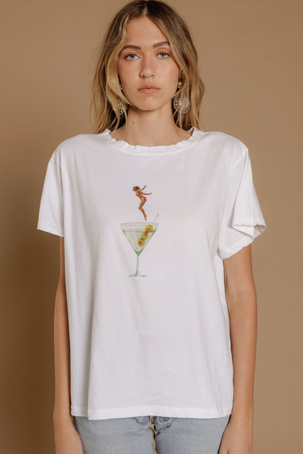 Person wearing a white t-shirt with a graphic design of a woman jumping into a martini glass on a brown background