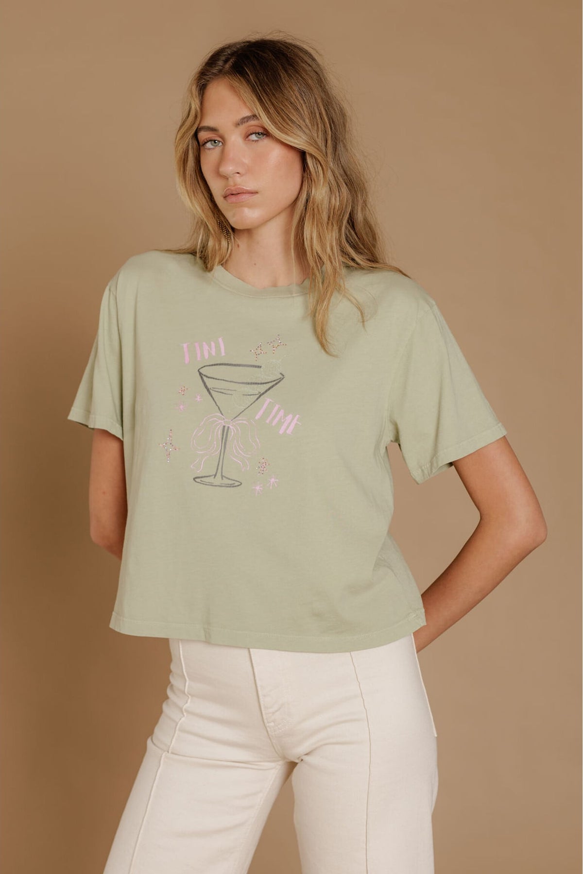 A woman modeling a pistachio green graphic t-shirt with a pink and gray martini glass design, paired with cream-colored pants, set against a solid tan studio background.
