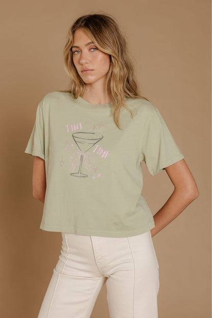 A woman modeling a pistachio green graphic t-shirt with a pink and gray martini glass design, paired with cream-colored pants, set against a solid tan studio background.
