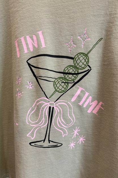 A close-up of a pistachio colored t-shirt featuring a graphic print of a martini glass with a pink bow, green olives on a skewer, and pink glitter-textured stars and the words 'FINI TIME'.