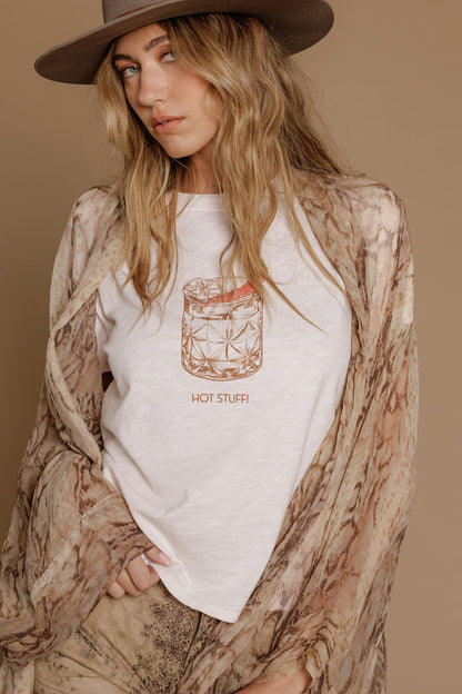 Woman wearing a powder pink t-shirt with a graphic design of a cocktail and text, standing against a beige background.