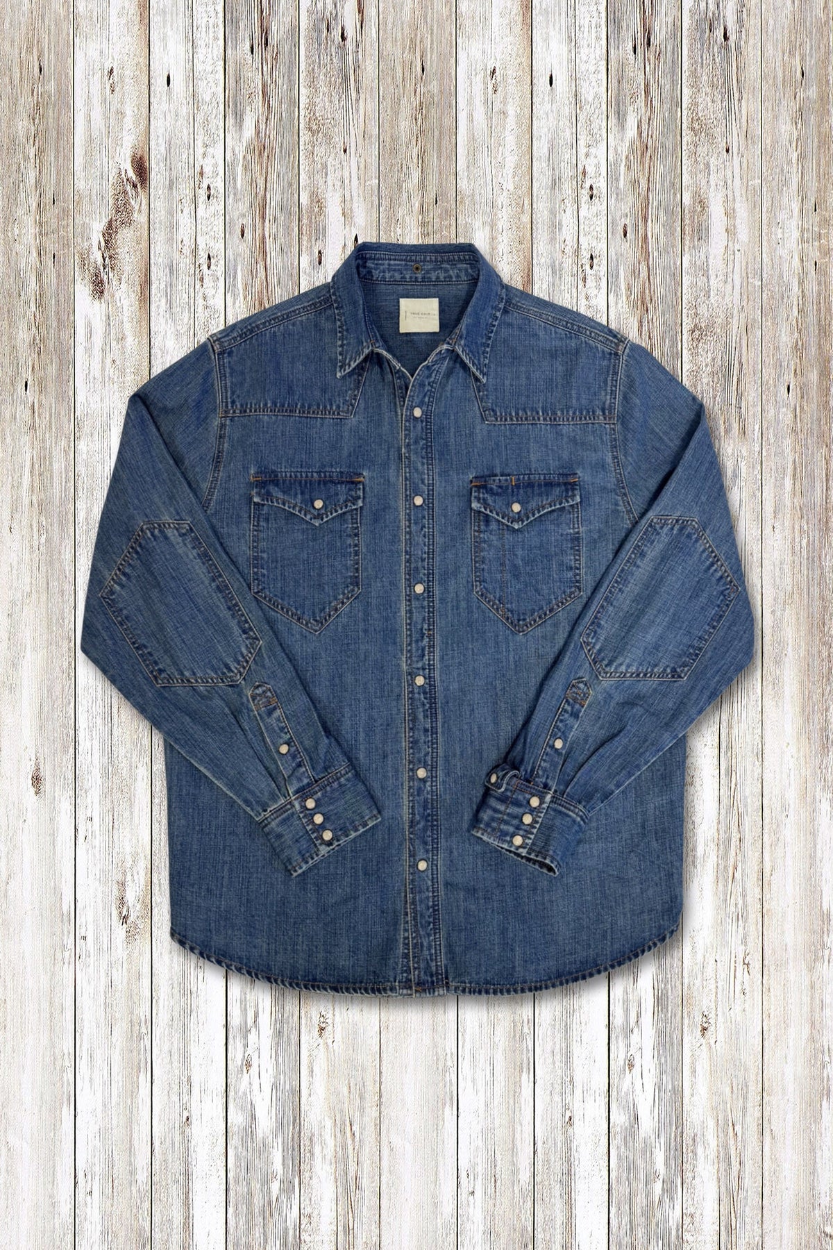 A blue denim long-sleeve western-style wayne shirt jacket with two chest pockets and buttoned cuffs, laid flat on a light wood plank background.