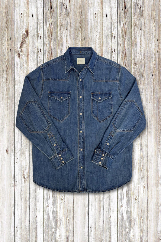 A blue denim long-sleeve western-style wayne shirt jacket with two chest pockets and buttoned cuffs, laid flat on a light wood plank background.