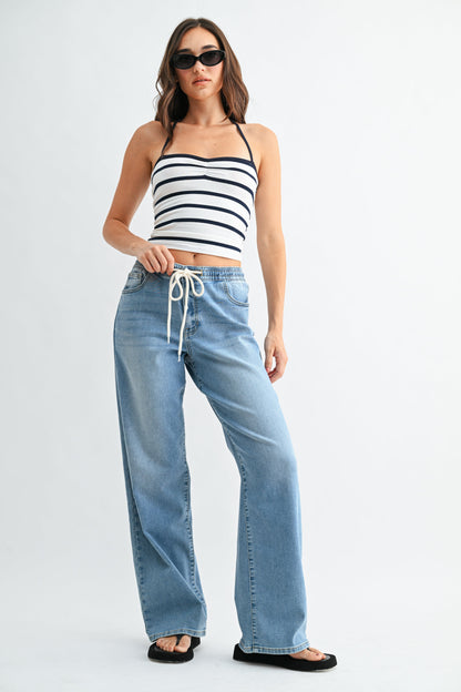 A woman modeling a black and white striped halter crop top paired with light-wash pull on drawstring wide-leg jeans and black flat sandals, standing against a plain white studio background.