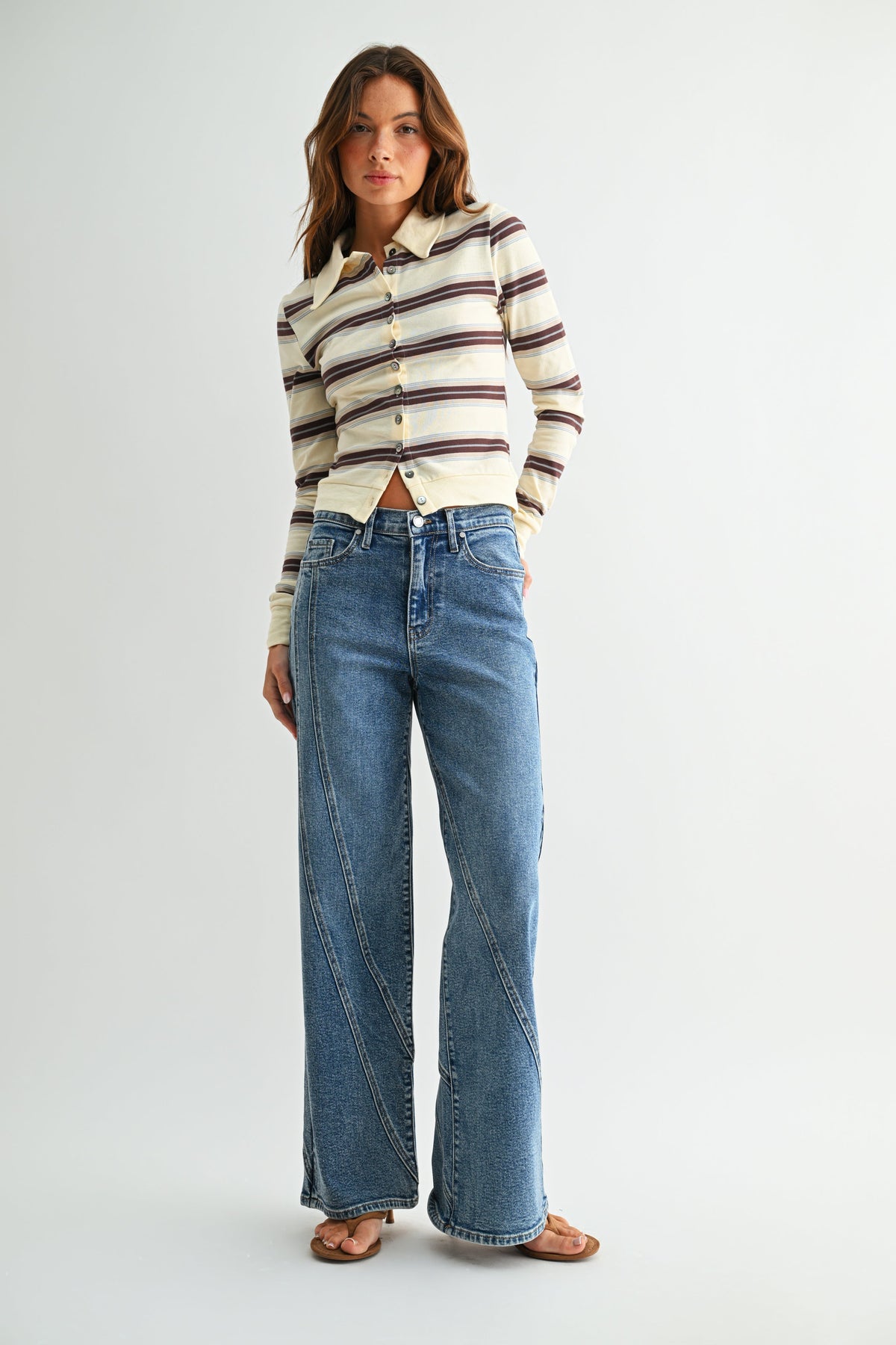 A woman modeling a cream long-sleeve button-up shirt with brown and dark stripes, paired with high-waisted, assymmetrical seamed straight leg blue jeans and brown flat sandals, against a light gray studio background.