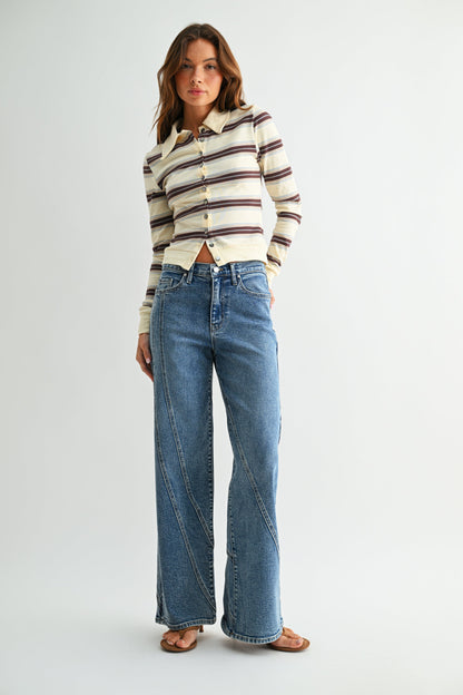 A woman modeling a cream long-sleeve button-up shirt with brown and dark stripes, paired with high-waisted, assymmetrical seamed straight leg blue jeans and brown flat sandals, against a light gray studio background.