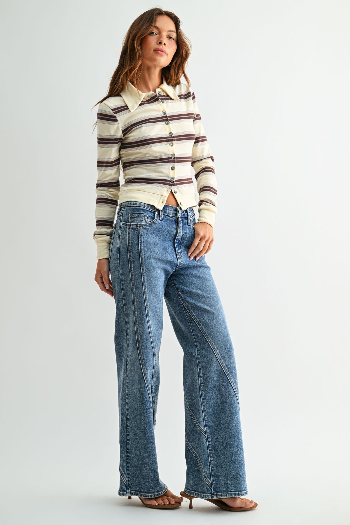 A woman modeling a cream long-sleeve button-up shirt with brown and dark stripes, paired with high-waisted, assymmetrical seamed straight leg blue jeans and brown flat sandals, against a light gray studio background.