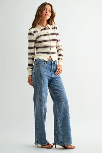 A woman modeling a cream long-sleeve button-up shirt with brown and dark stripes, paired with high-waisted, assymmetrical seamed straight leg blue jeans and brown flat sandals, against a light gray studio background.
